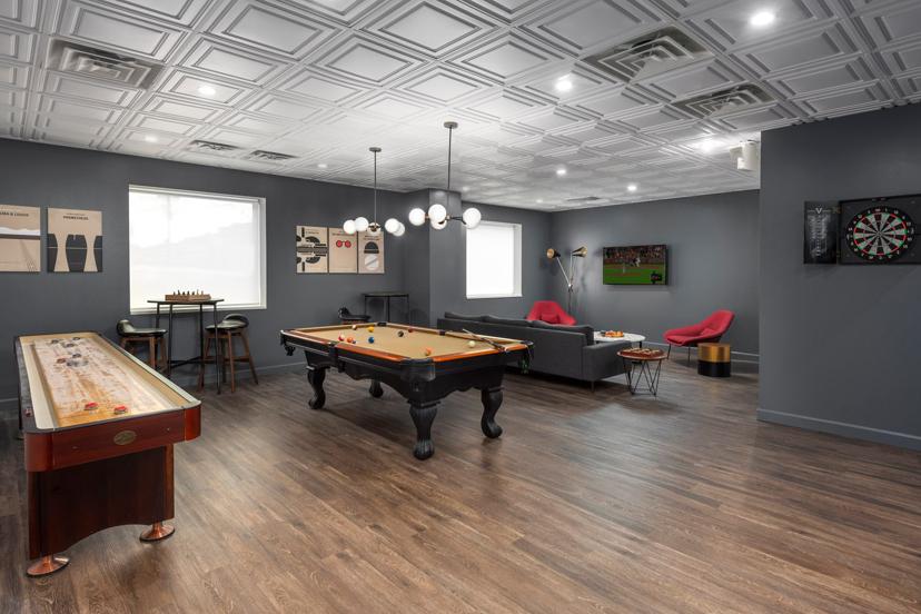 Modern game room with a pool table, shuffleboard, dartboard, bar stools, and a TV. Gray walls, wooden floor, and stylish lighting.