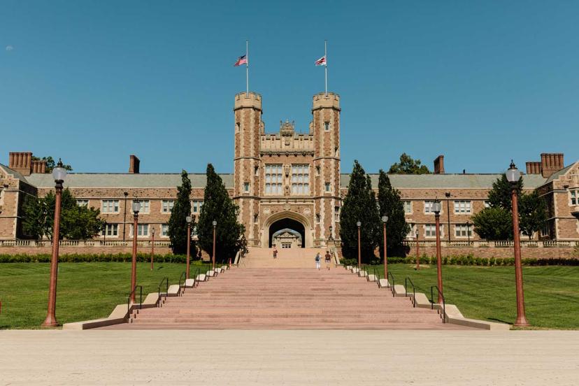 A large brick university building with twin towers, flags flying, and a grand staircase leading up, set against a clear blue sky.