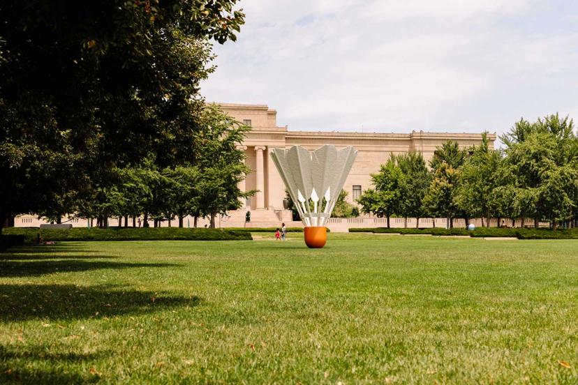 A giant shuttlecock sculpture stands on a green lawn with a classical building and trees in the background.