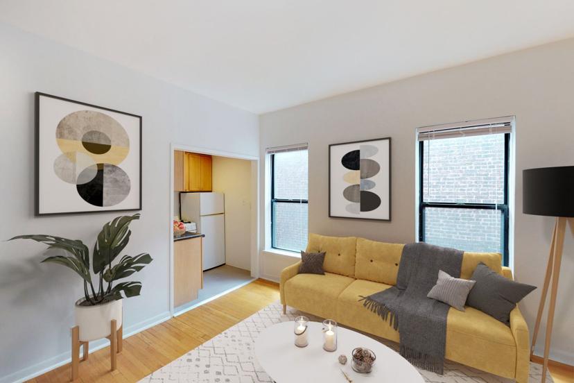 A cozy living room with a yellow sofa, abstract wall art, potted plant, and a kitchen in the background. Candles and decor on a white table.