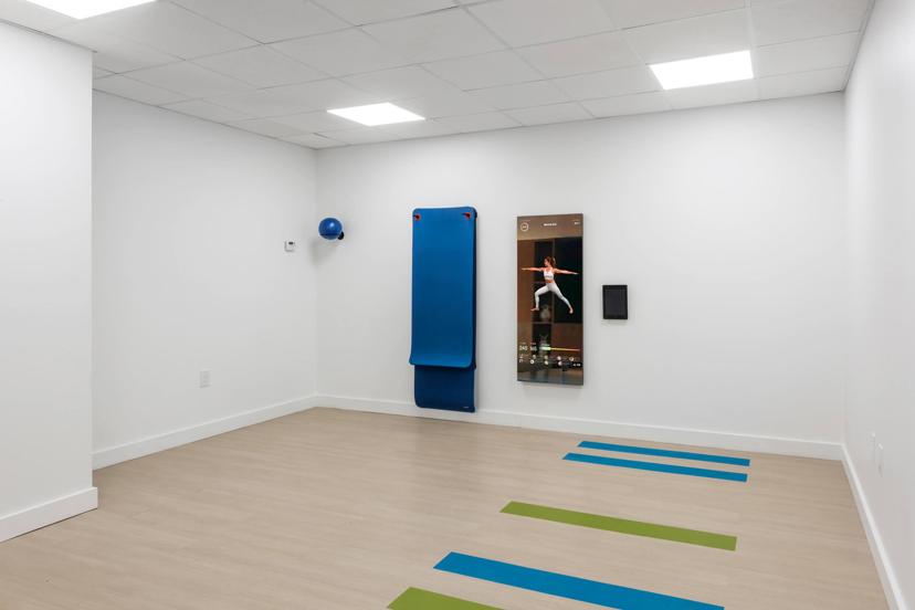 A minimalist gym room with a smart fitness mirror, blue mat on the wall, blue and green stripes on the floor, and a blue yoga ball.