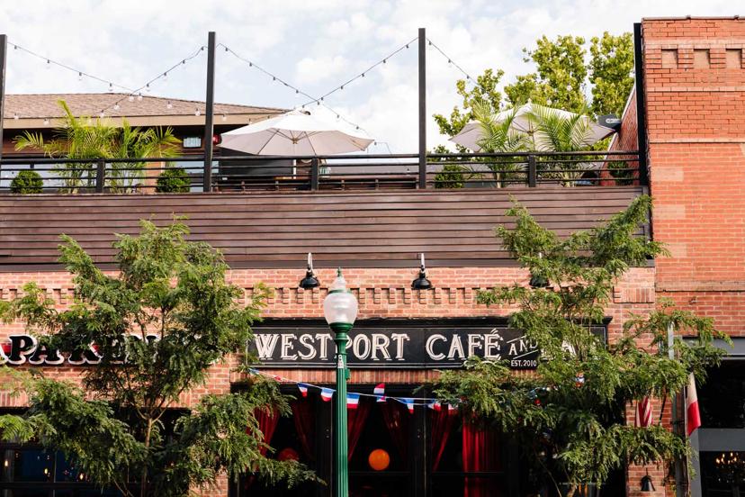 Brick café exterior with "Westport Café" sign, trees, string lights above, and a patio with umbrellas in daylight.