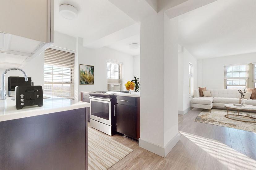 Modern open-plan kitchen and living area with white walls, dark cabinets, and light wood flooring. Natural light from large windows brightens the space.