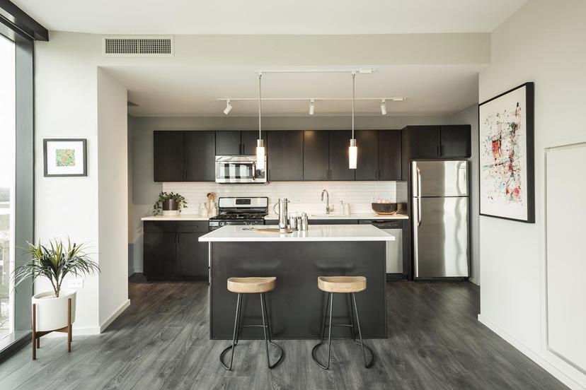 Modern kitchen with dark cabinets, stainless steel appliances, white island, two wooden barstools, and decorative plants and artwork.