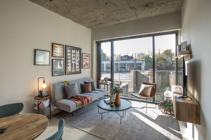 Modern living room with a gray sofa, decorative pillows, and wall art. Large windows let in natural light. A round coffee table and seating area are present.