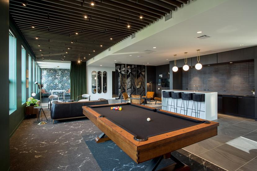 Modern lounge with a pool table, bar area, and seating. Stylish decor includes black and white wall art, green curtains, and pendant lights.