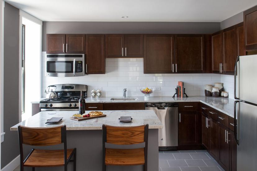 Modern kitchen with dark wood cabinets, white tile backsplash, and marble countertops. Stainless steel appliances and two wooden chairs at the counter.