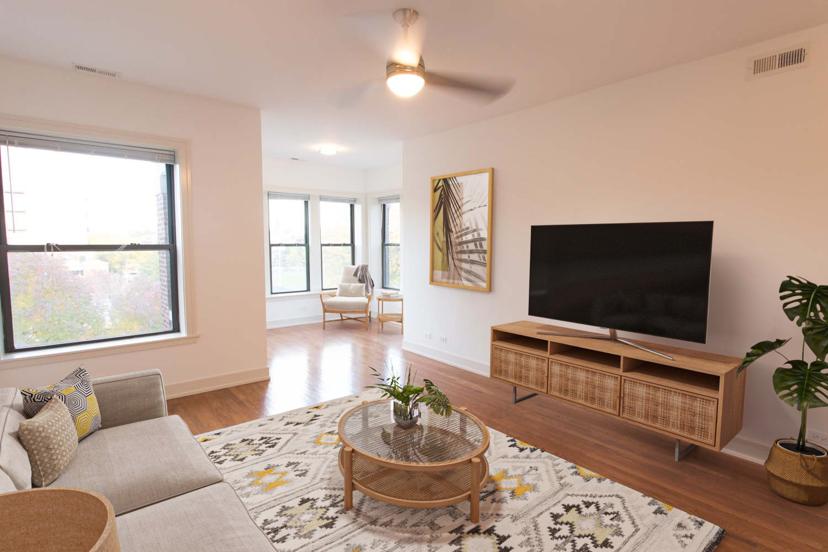 Modern living room with a beige sofa, patterned rug, glass coffee table, large TV on a wood stand, potted plant, and large windows with natural light.
