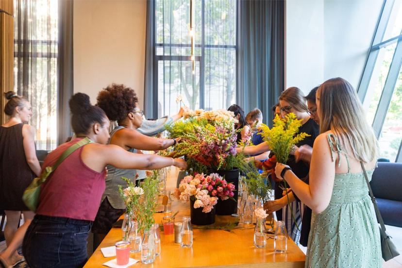 People arranging colorful flowers at a long wooden table, surrounded by glass jars and natural light from large windows.
