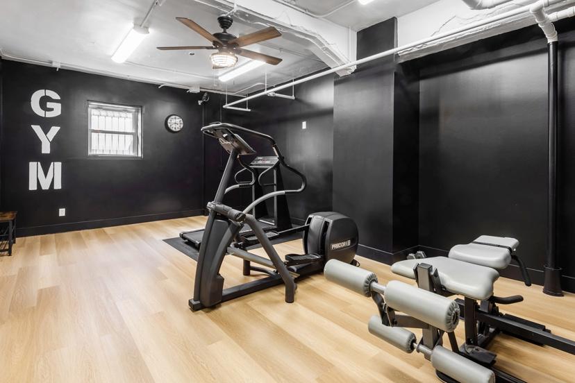 A small home gym with black walls, featuring a treadmill, adjustable bench, weights, wooden flooring, ceiling fan, and a wall clock.