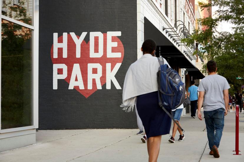 People walking past a mural with "HYDE PARK" in bold white letters on a red heart, painted on a brick wall in an urban setting.