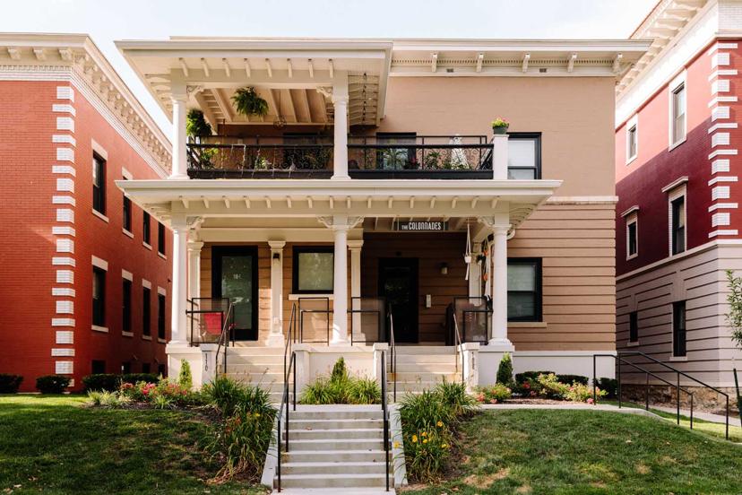 A two-story brick building with a white columned porch and balcony, surrounded by well-maintained grass and plants.