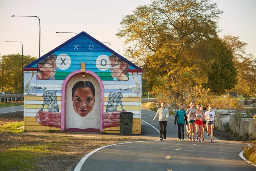 A group of women jog toward a colorful, graffiti-covered building, featuring large portraits, on a sunny day with trees in the background.