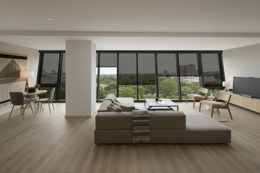 Modern living room with large floor-to-ceiling windows, a gray sectional sofa, TV, dining area, and light wood floors, overlooking a cityscape.