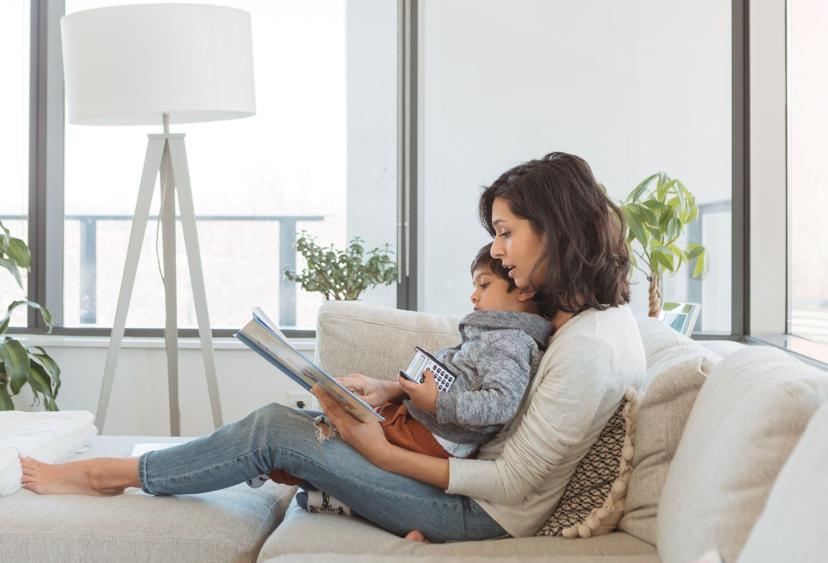 A woman sits on a couch reading a book with a young child on her lap, surrounded by bright windows and plants.