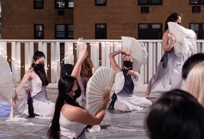 Dancers perform with large white fans on a rooftop, wearing masks and flowing white and gray costumes, with string lights in the background.
