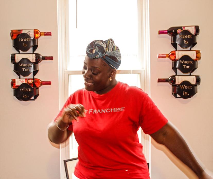 A person in a red shirt smiles and gestures in front of a wine rack that reads "Home Is Where The Wine Is," with wine bottles displayed.