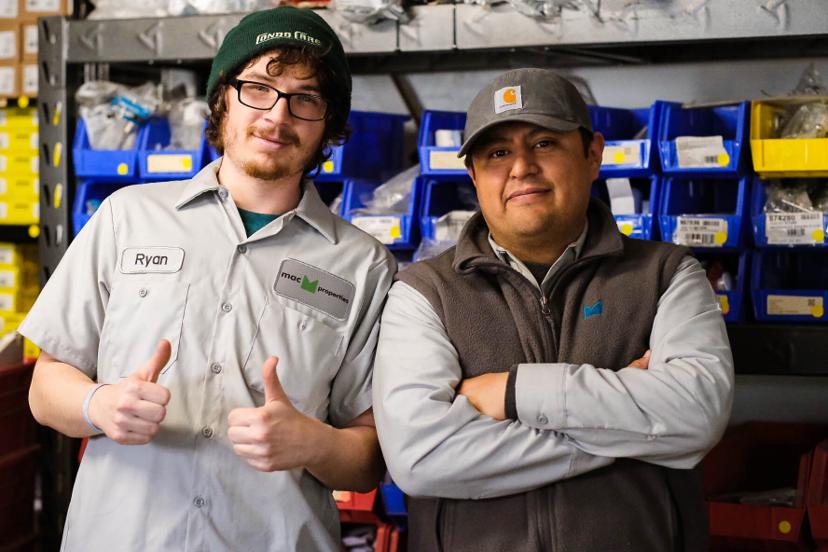Two men in work uniforms stand in a warehouse, one smiling with thumbs up and the other with arms crossed, surrounded by shelves.