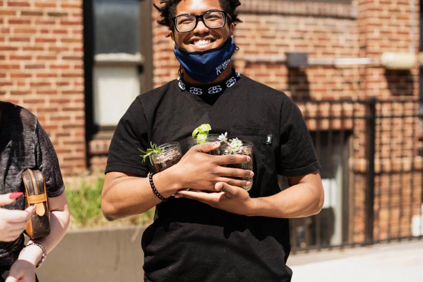 A person in a black shirt smiles while holding jars with small potted plants outside a brick building.