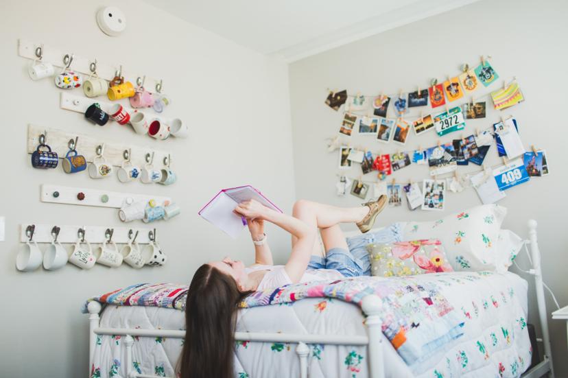 A person lies on a bed reading a notebook in a cozy room with wall art and mugs hanging on hooks.