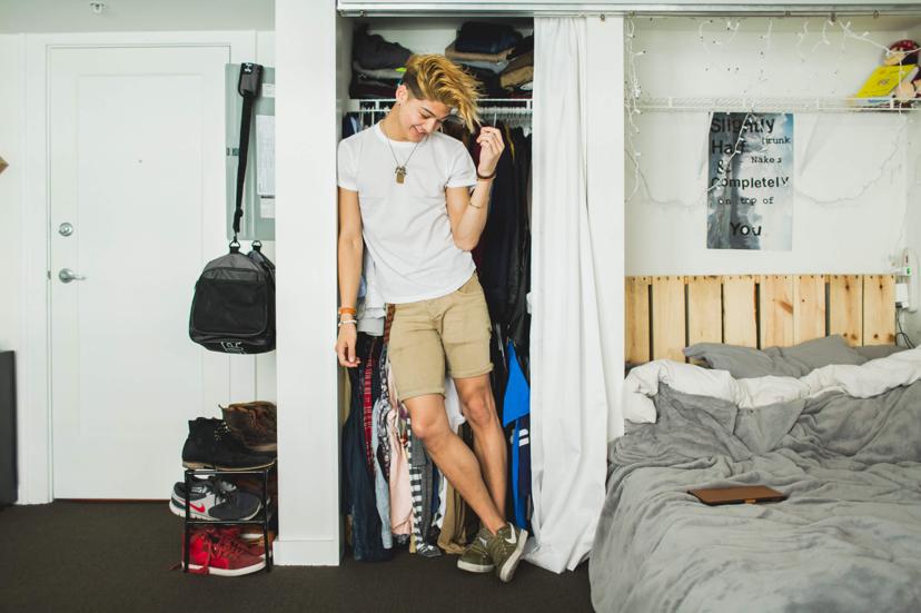 A person stands in a closet wearing a white shirt and khaki shorts, smiling. The room has a bed with gray bedding, shoes, and a bag on the floor.