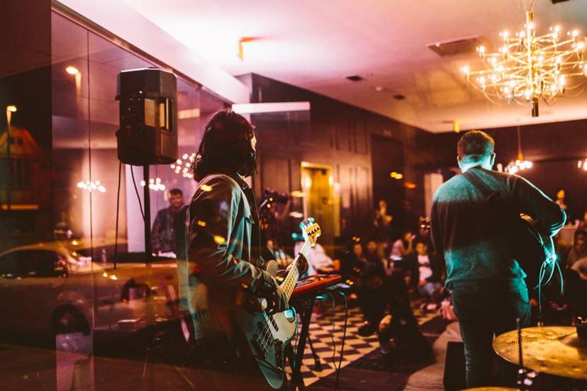 Musicians perform in a cozy, warmly lit venue with an audience seated on the floor. A chandelier and reflections in glass add ambiance.